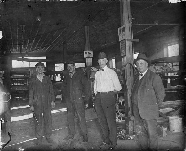 Five men standing in the J.W. Meiklejohn & Son's Prison City Garage. The men on the right are wearing business clothes and hats, the men on the left are wearing mechanic's coveralls and caps. A fifth man is partially out of the image on the left, wearing a suit. The garage is filled with automobiles and tools. Light is streaming in the window and a large door. Several notice and policy signs are tacked to the wooden posts. One reads: "There is only one price on these cars, The List Price." 