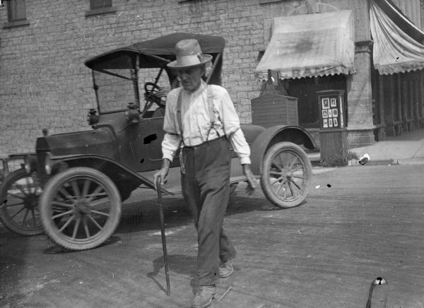 A man walks across the street with the aid of a cane. An Model T style automobile turns the corner behind him. In the background is a business, the awning reads: "A. Rank." An advertising kiosk displays photographic portraits.