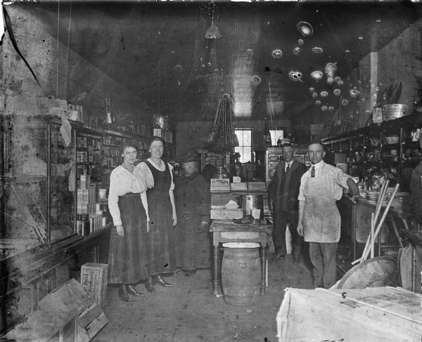 Three woman on the left and two men on the right are standing in a general store. Shelves line the wall and a display table is in the center with a covered barrel. Some of the products on display are, Cracker Jacks, Bob White laundry soap, dishware, produce from Libby, McNeil & Libby, and many unidentified boxes and bottles. Brooms hang from the ceiling.