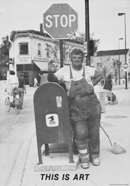 Poster of Art Nesson, a well-known figure on State Street. Art made his living as a window washer, dishwasher and manual laborer. He is leaning on a mailbox along West Gilman Street. There is a broom propped up behind him and he is waving to the camera. Behind him is a "Stop" and a "One Way" sign at the corner. Pedestrians, including a man riding a bike, and a woman pushing a stroller, are on the street and sidewalks. Across the street is the Chocolate Shoppe Ice Cream Store on State Street. Text at the foot reads: "THIS IS ART."