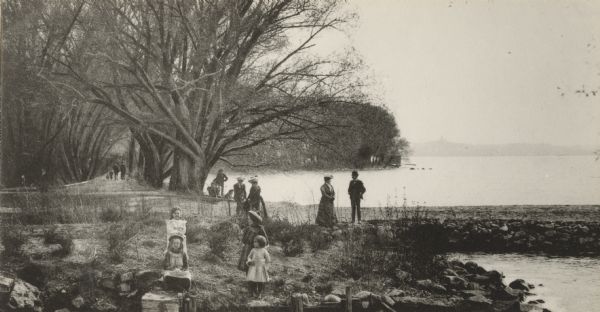 A view across Lake Mendota from Tenney Park. A group of four children are on the near shoreline. A group of adults are standing and sitting in the midground, and further down on the left another group is walking down the path along the lake under the trees. On the right is the breakwater. In the far distance is the shoreline of the isthmus and a silhouette of Bascom Hill.