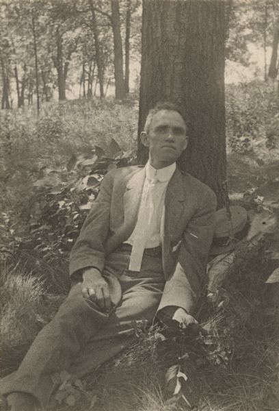 Portrait of Charles Brown. He is sitting outdoors on the grass leaning against a tree trunk and holding a plant in his left hand.