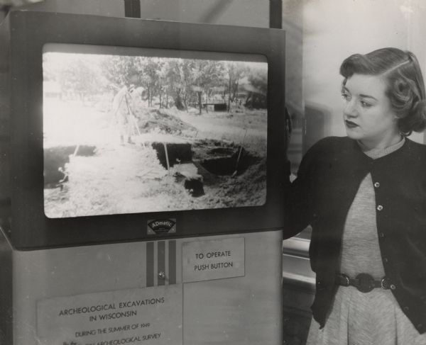 Shirley Jeffrey standing and watching an automatic projector which accompanied an exhibit. The projector was adapted by Harry Lichter of the museum staff. The sign below the projector reads: "Archeological Excavations in Wisconsin during the summer of 1949."