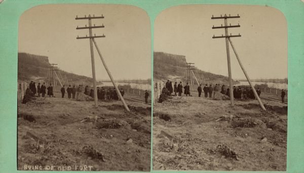 Stereograph of fireplace ruins on the site of an old French fort, possibly built by Nicholas Perrot. There is an electric pole behind the fireplace and a group of men and women are in the background. Railroad tracks are along the shoreline of what may be the Mississippi River on the right.