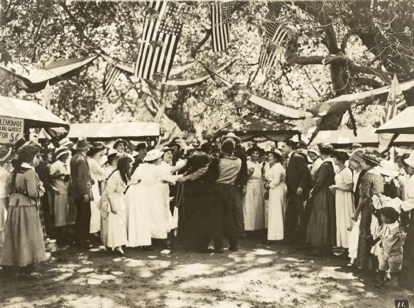 Film still exterior of a group of people at a picnic. A man is holding a bear with a chain in the center of the image. Booths can be seen in background, one with a sign offering glasses of lemonade for five cents.