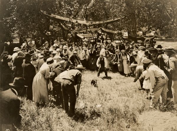Film still of a group outdoors at a Fourth of July picnic. A man is chasing a small pig in the center of a group of people. 


