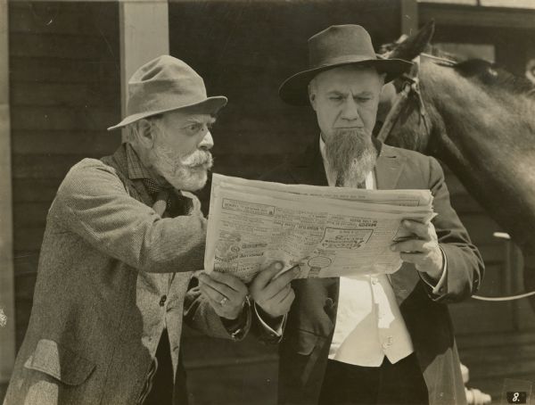 Film still of two men standing outdoors looking at a newspaper. A horse is tethered behind them.