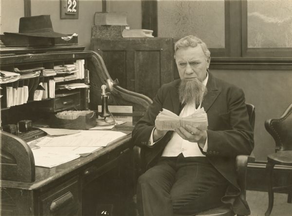 Film still of a man seated at a roll-top desk in his office.
