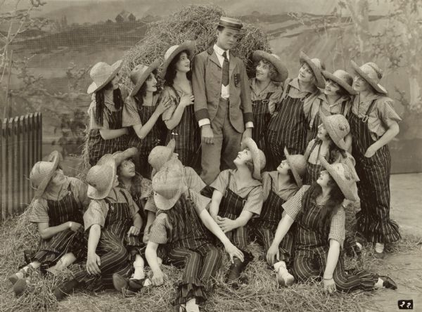 Film still of a male actor in a stage musical surrounded by actresses in matching costumes.
