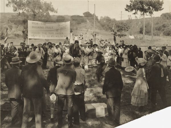 Film still of a picnic in "The Clod Hopper". Spectators are at a mule-riding attraction. There is a man on a mule in a enclosure, while a second man in a cowboy hat holds a rope attached to the mule. Sign reads "Ride the Bucking Mule."