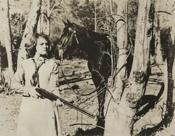 Actress Josie Sedgwick plays Betty Clayton in this western. Betty is in charge of the Lazy Y ranch, and she is friendly with Calumet Marston, whose father Jim had put her in charge of the ranch while Clayton was absent. In the story Betty uses her rifle to wound Tom Taggart, a squatter, when he shoots Clayton in an ambush. In this image, Betty is standing in the woods near her horse, looking out warily with her rifle.