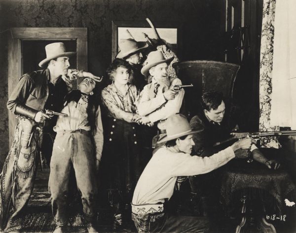 Actress Josie Sedgwick as Betty Clayton and six other men, some with guns, holed up in a ranch house, shooting toward a window. One of the men stands with arm across his forehead, and another man holds Betty by the wrist. 
