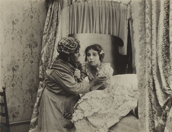 Film still of Interior bedroom with young woman in bed being comforted by a servant - a white actress in blackface. ("Black face" is written on the back of the Still.)  Four-poster-type bed and decorative wallpaper are visible.