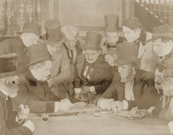 Film still of group of well-dressed men at a table playing a game of cards; money and drinks are on the table. The men are all wearing top hats.

