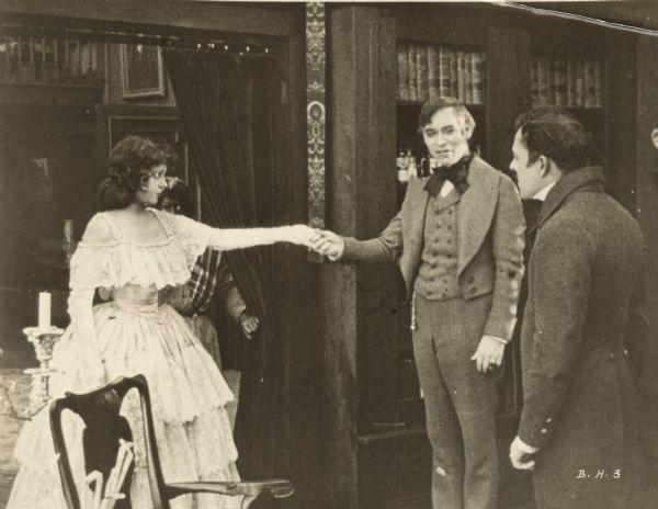 Film still of two women, one extending her hand to a man, another man is standing by; a female servant in blackface is in the background.


