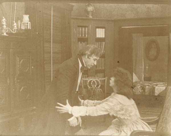 Film still of interior with a man standing and a woman sitting in front of him with her hands reaching out and touching his elbows. She is speaking earnestly to him.