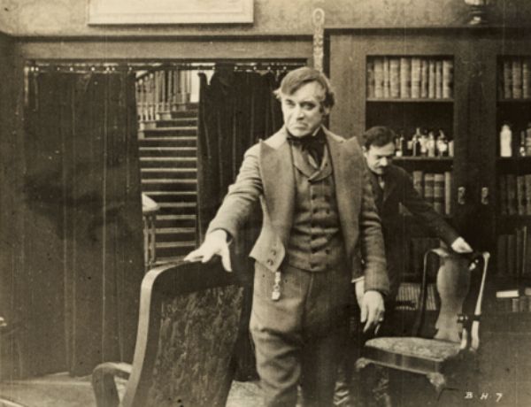 Film still of an interior with two men, one in foreground, well-dressed, right arm resting on back of chair.