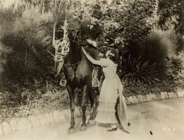 Film still of an exterior with a man on a horse speaking with a woman standing below, reaching up to him.
