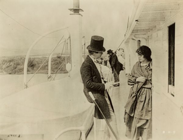 Film still of an exterior with a man and woman in conversation on a riverboat deck. Other people in background. 


