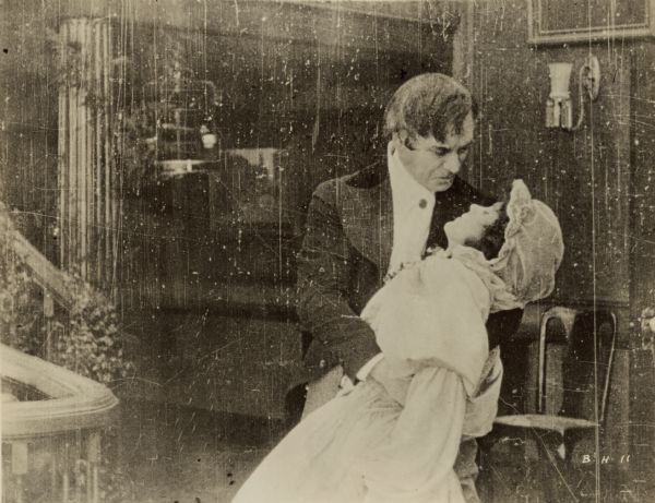 Film still of an interior, man and woman. He is embracing her as she leans back with her eyes closed.

