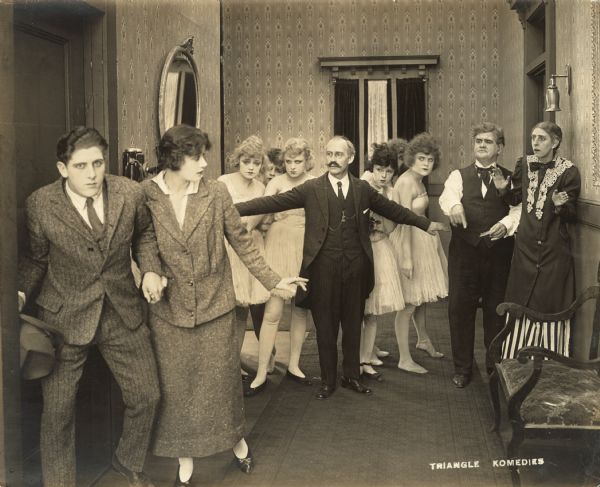 Film still depicting a group of men and women, some of the women dressed in gown costumes.  
