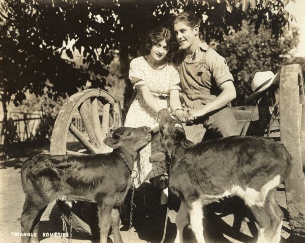 Film still - exterior shot with actor and actress holding hands as they feed two calves.  
 	


