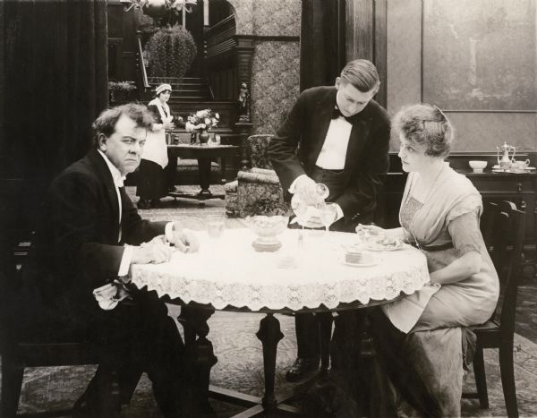 The millionaire David Waltham and his wife (played by George Fawcett and Louise Brownell) are being served at a table by their butler while a maid arranges flowers in the background in a scene still for the 1916 silent drama "The Corner."