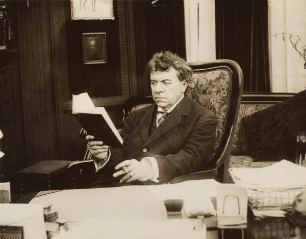 The millionaire David Waltham (played by George Fawcett) sits at his desk reading a book, smoking, a cigar, and scowling in a scene still for the 1916 drama "The Corner."