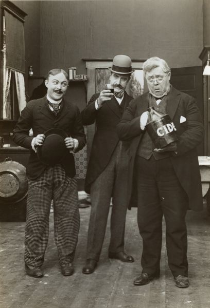 Film still - interior with three actors and a jug of cider, one holding a glass in a toast.


