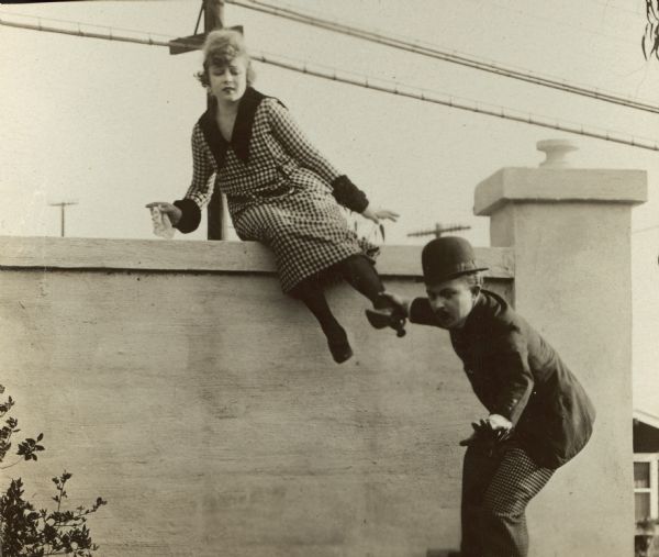 Film still - exterior with an actress climbing over a cement or plaster wall. Actor below in foreground has a grip on her left ankle.



