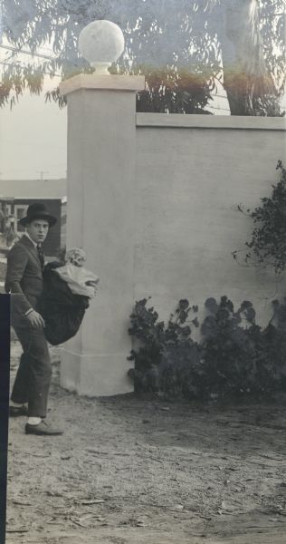 Film still - exterior with a man standing near a high wall of plaster or cement, holding a large bag of props.


