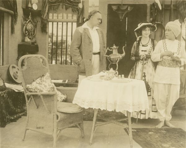 Film still of interior scene, exotic atmosphere, with actor and two actresses, one in Indian costume with turban.