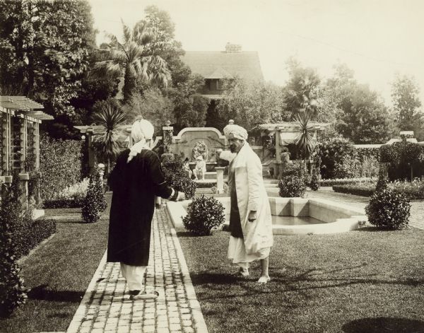 Film still of exterior garden with two actors in Indian costume with turbans in conversation.

