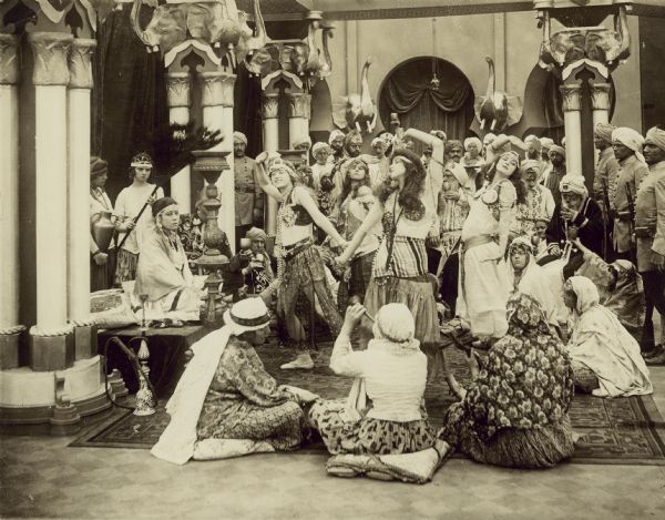 Film still of interior palatial scene with large group of actors and actresses dancing and celebrating in Indian costumes, some with turbans.
