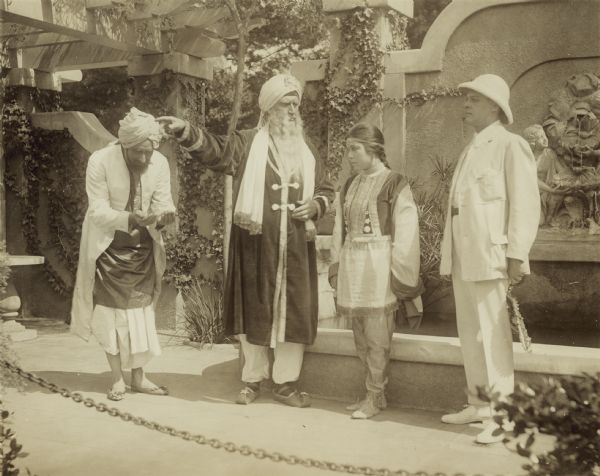 Film still of exterior garden with three actors, two in Indian costume with turbans. One actress in Indian costume. One actor in "colonial" costume.