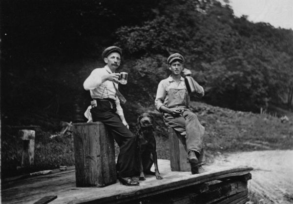 Two men are drinking, one from a mug and one from a bottle near the Cassville Brewery. The man on the left is Frank Feiker, and the photograph is taken with a self timer. They are sitting on boxes on a platform outdoors wearing caps and casual clothes. The dog sits between the two men.