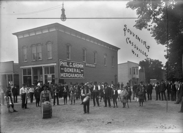 Town members surround musicians posing with their instruments. They are all standing in the street in front of the General Merchandise store. A Frank Feiker 'Souvenir' postcard. A building in the background has a sign that reads: "Dr. Feiker."