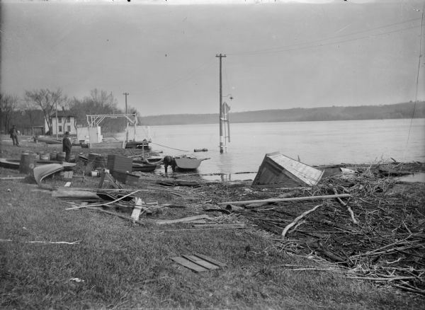 View along shoreline of man examining a rowboat while crouching in the water wearing boots. Behind him is a ferry. There are several men on the shore watching. Debris litters the shoreline, and power lines are standing in water. 
