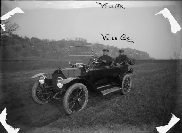 Two men sitting in an open car in a field. There is a bluff in the background. On the front of the car underneath the windshield is a license plate with the year 1914. Feiker has labeled the photograph "Veile Car," an alternate spelling of the family name.