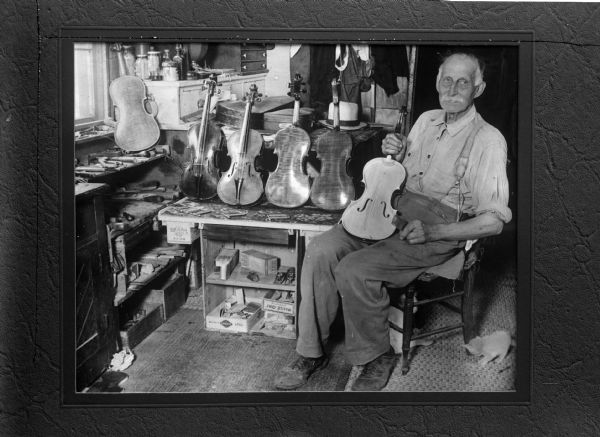 A man sits in his shop, holding a violin. Five more violins are arranged next to him. Caption with original negative reads "Old-time fiddle-maker in his shop." The man is John Graybill, a woodworker from Cassville who created violins throughout his lifetime.