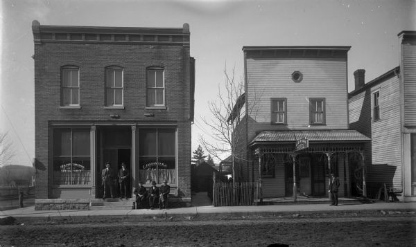 View across dirt road of two businesses. On the left is a two-story brick building with large windows which are painted with signs that say: "Habermann Buffet" and "Liquors & Cigars." Five men are posing in front, with two standing in the doorway and three others on a bench on the sidewalk. A young boy or man is standing in front of the Habermann Hotel, which is a clapboard building on the right with a front porch and a false front.