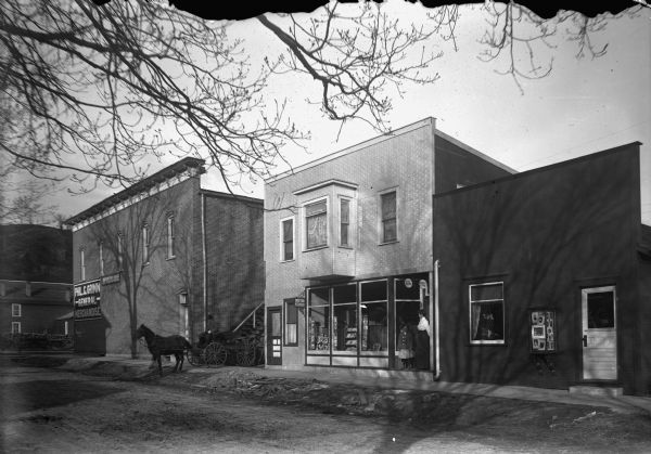 Three storefronts on a dirt road. A man, said to be Charles Grimm, is driving a horse and buggy. The building in the center is Feiker's photo studio. Near a door is a glass case display of photographs mounted on an exterior wall just above the sidewalk. A woman and young girl stand in a doorway near show windows with a sign that reads: "Doctor Feiker". On the far left at the corner is Phil. C. Grimm General Merchandise.