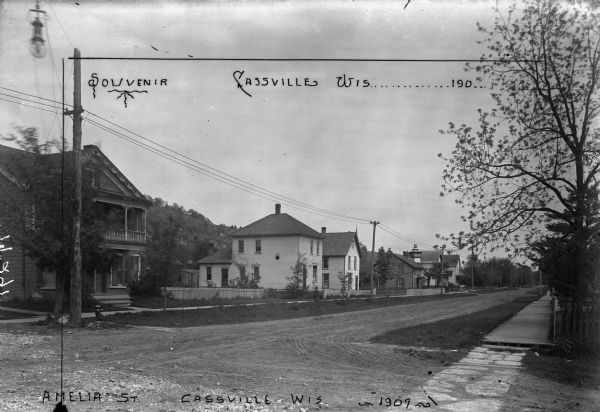 View across unpaved street of several houses, with board sidewalks, fences and power lines. In the background is a bluff.