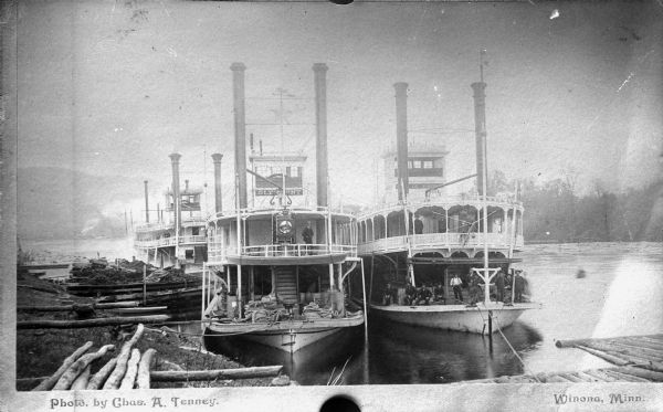 View from shoreline of three steamboats at dock. Men are sitting and standing on the boats. Partial names of two of the boats are visible.