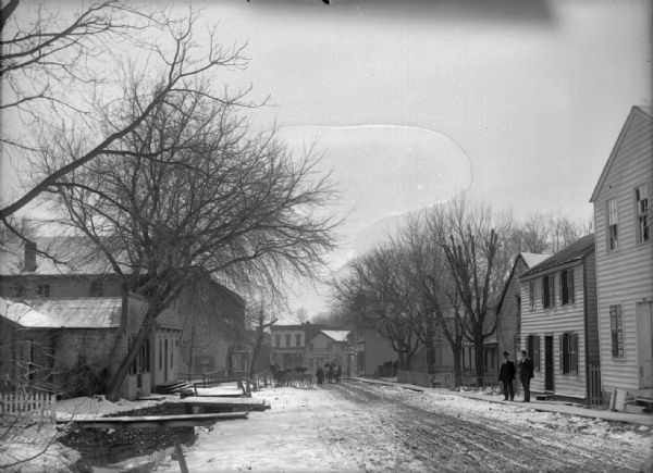 An unpaved Potosi street in winter. There are unidentified men standing in front of a building, and in the distance are carriages with horses.