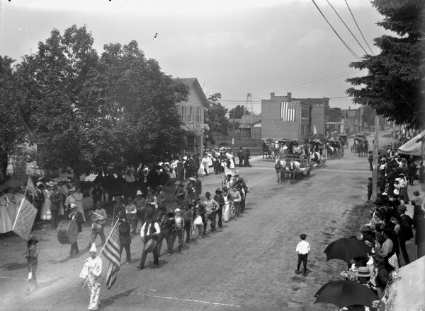 Elevated view of parade with a band of clowns. One young boy dressed as a clown at the front is holding the American flag, and another clown holds a sign that says: "Don't judge us by our looks but by our music." They are followed by other clowns holding musical instruments. Crowds line the sidewalks to watch. On the street in the background are horse-drawn wagons and carriages.