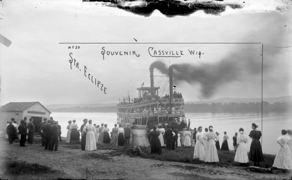 A Frank Feiker 'Souvenir' photograph, labeled 'No 20' in the upper left corner. View from shoreline of a steamboat full of people arriving. There is a large group of men and women standing on the shore. A small building on the left says "Eclipse Company."