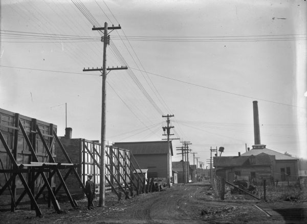 A man stands near the backs of billboards on the left along a street or alley. Power lines lead to the horizon, over what appear to be businesses or industrial buildings. A building on the right has a large smokestack rising out of the middle of the roof.