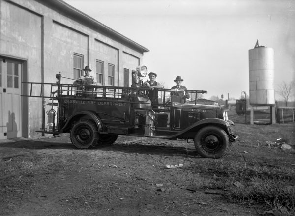 Three men, Henry Dietrich, Frank Eckstein and Peter Bausch, are posing with the fire truck they built. They took the truck to Dubuque to have the bright work done. There is a large water tank in the background to the right. The fire truck says: 'Cassville Fire Department, C.F.D.'