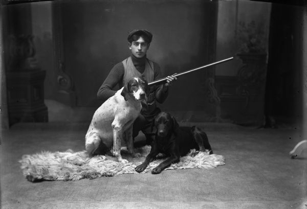 Studio portrait in front of a painted backdrop of a young man holding a shotgun, and two dogs posing on a sheepskin rug.